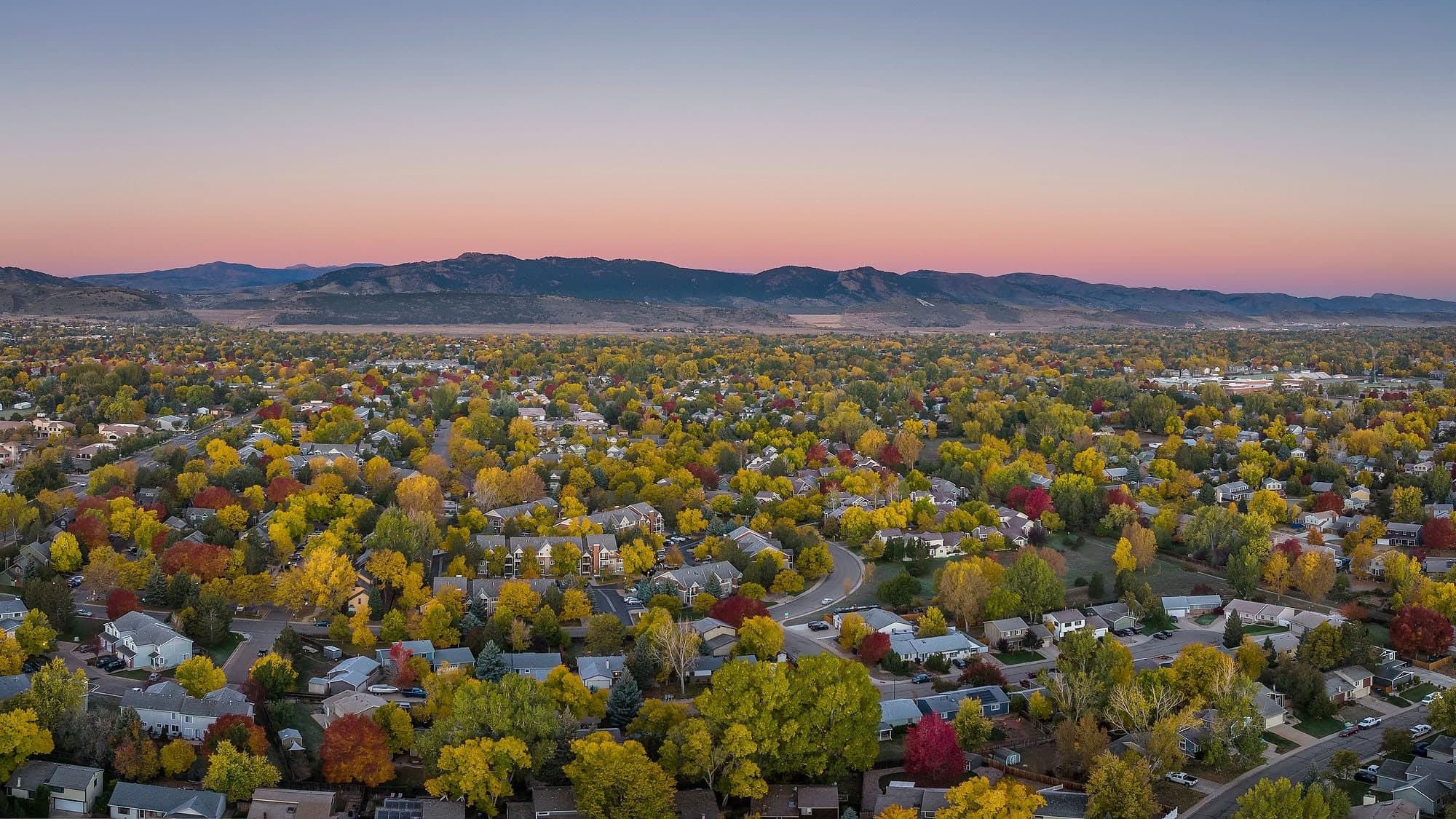 Fort Collins from the air at dusk: neighborhood rooftops among gold and red fall trees, with the foothills along the horizon under a pink sky.